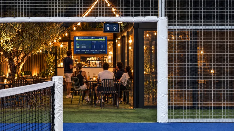Un tablet su un supporto a collo d’oca all’angolo superiore dell’ingresso del campo; la stessa partita appare sulla TV del bar, seguita dalle persone ai tavoli
